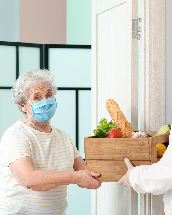 An older adult standing in doorway, with mask on accepts a box of food.