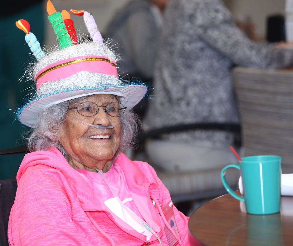Photo of a smiling older adult wearing a birthday cake hat on their head, and using supplemental oxygen.
