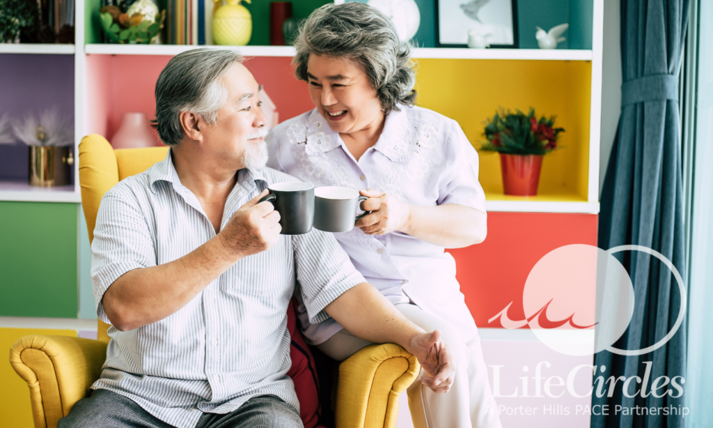 Older man and woman sit together, clinking coffee cups in front of bright shelving in their home. 