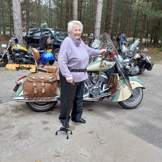 A woman with short white hair, and a walking cane stands next to a motorcycle.