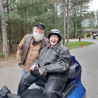 A person with a white beard stands behind a smiling older person sitting on a blue motorcycle. 